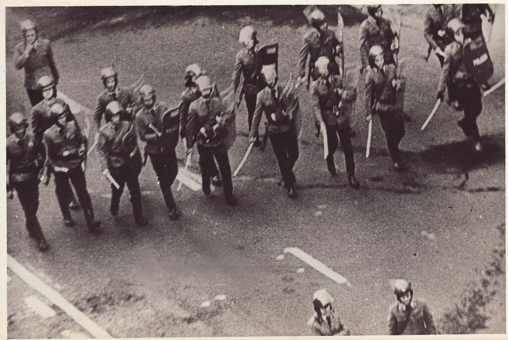 Grupa zomowców idzie w szeregu z pałkami i tarczami