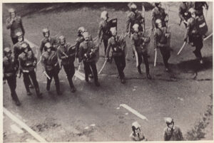 Grupa zomowców idzie w szeregu z pałkami i tarczami