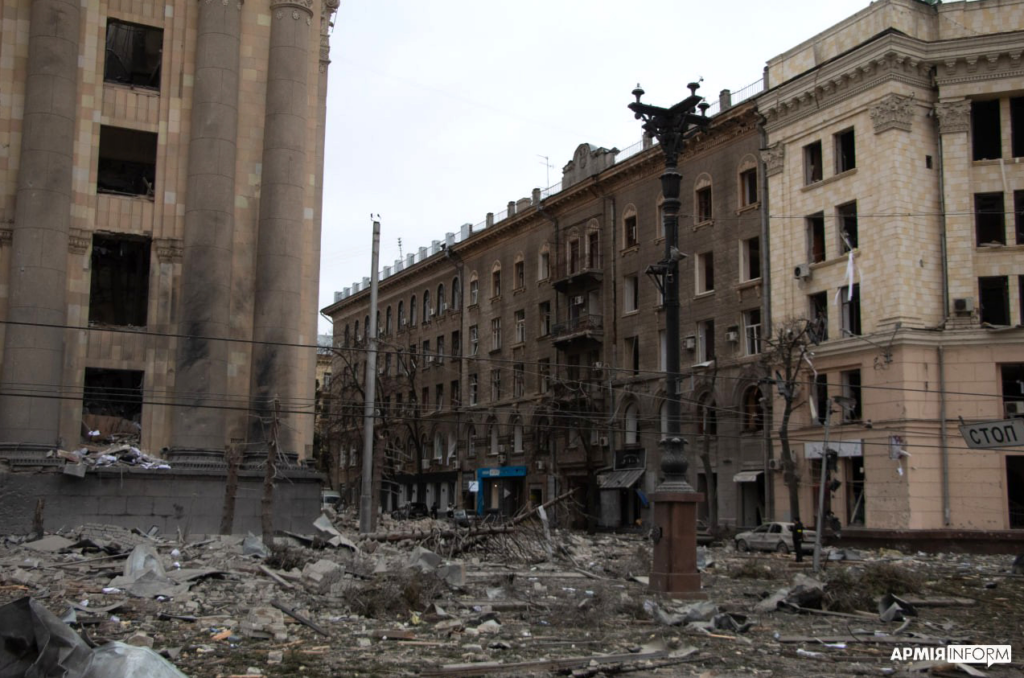 Widoczne dwa budynki. Jeden w całości. Budynek zniszczony, nie ma w nim okien, są ślady po pożarach. Na ulicy dużo gruzu.