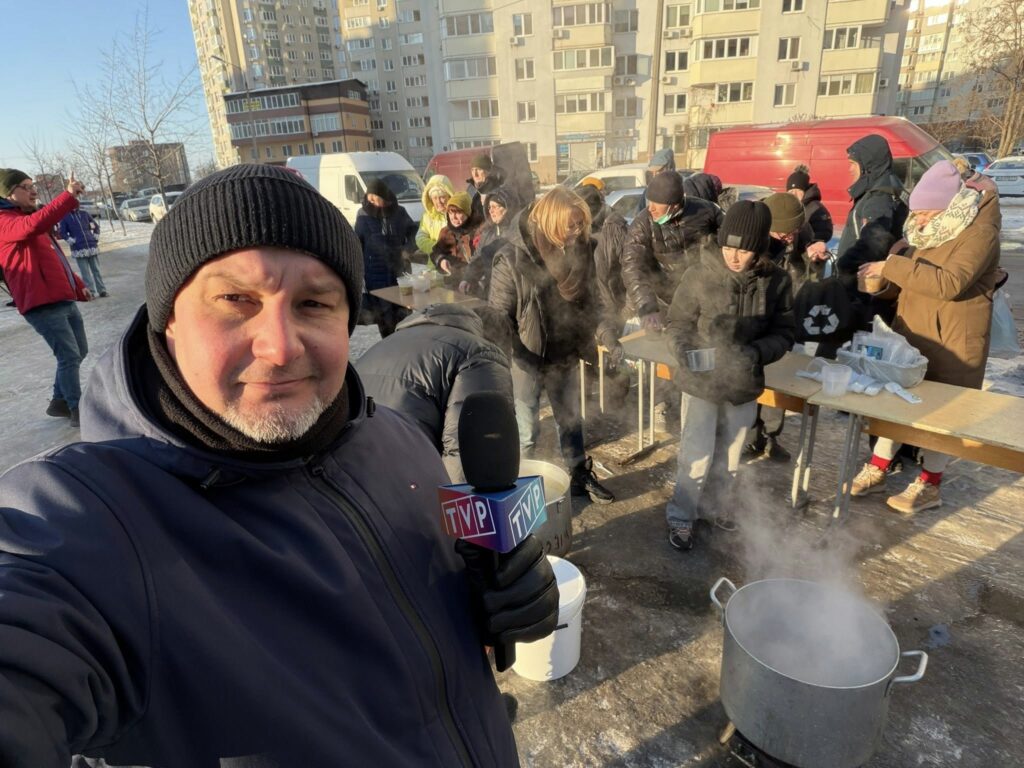 Człowiek w czapce robi sobie selfi. W tel stoły, na których stoją produkty żywnościowe. Są rozdawane. Obok mężczyzny kociołek z czymś gorącym. Paruje. Wszyscy ciepło ubrani. Na zdjęciu około 15 osób.