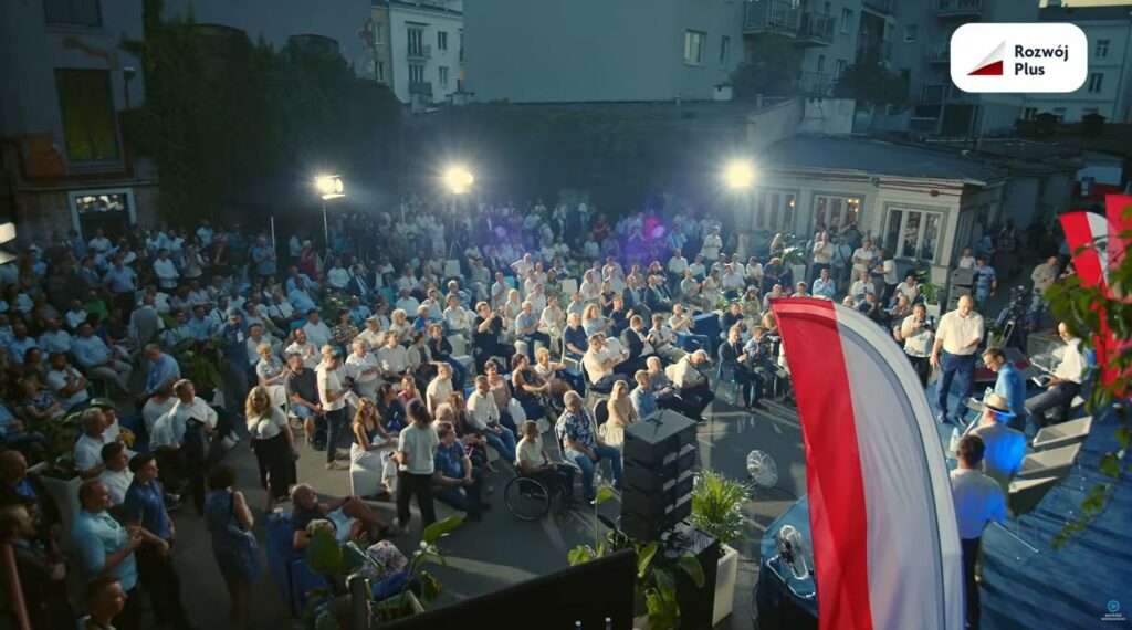 Widok ludzi siedzących naprzeciwko sceny. Jest ich około 200. Na pierwszym planie flaga biało-czerwona