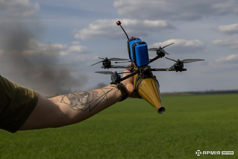 ręka trzyma drona FPV, pod którym podwieszony jest pocisk artyleryjski
